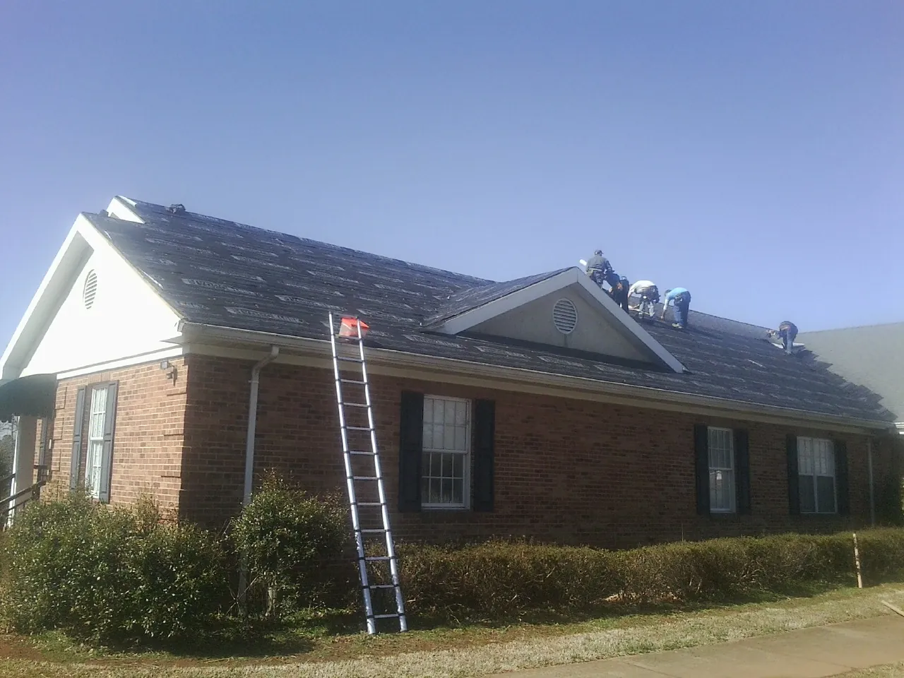 Crew tearing shingles off a brick ranch roof with a ladder set — Metal Roof Repair work by our crew in Piffard