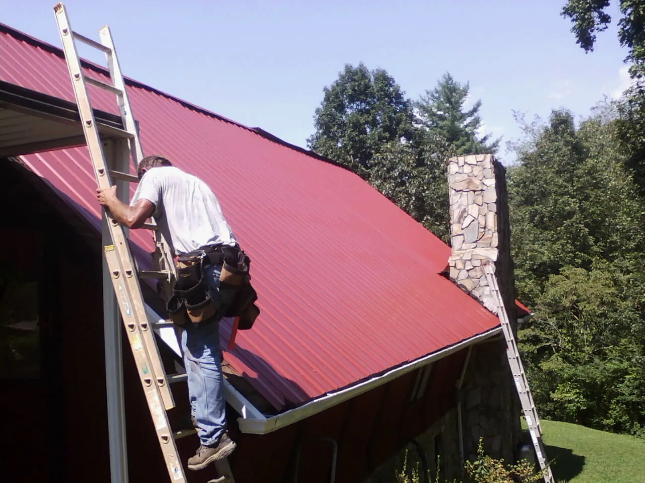 Roofer on a ladder beside a red metal roof and stone chimney — Soffit & Fascia Repair by Roofing Brothers in Piffard
